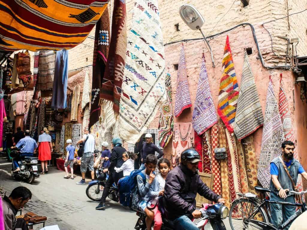 Tourists walking and people riding around on bikes through the Medina with rugs hanging on a wall in the background on a sunny day as motorbikes drive through Marrakech. You can see people driving in morocco here.