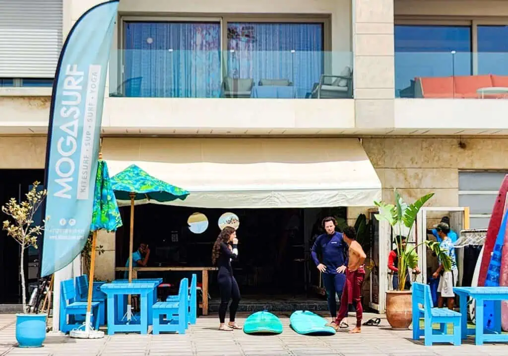 View of the exterior of Moga Surf Shop in Essaouira. You can see people out front and blue/green boards on the ground.