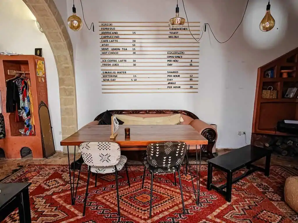 View of the work space in NOQTA with a menu on the wall for pricing and the cost of drinks. You can see a vibrant ruge and a couch against the wall with two chairs and a table in front of the couch. This is a great place for coworking in essaouira.