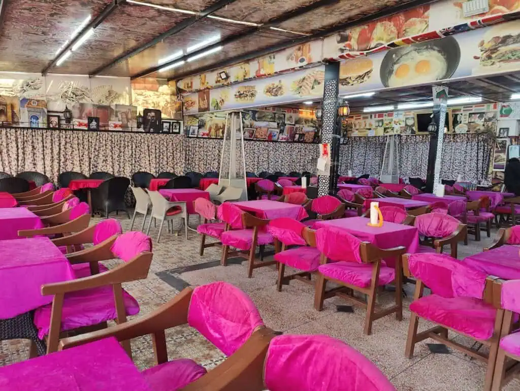 A view of the inside of Restaurant Atlas in Sidi Mokhtar. You can see tables with pink cushions and tables clothes beneath walls lined with pictures of food. You will stop here when you take the bus from Marrakech to Essaouira.