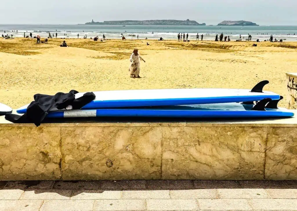 View of two blue surfboards sitting upside down on a wall in Essaouira. You can see the beach in the background and people walking on the beach on a sunny day.