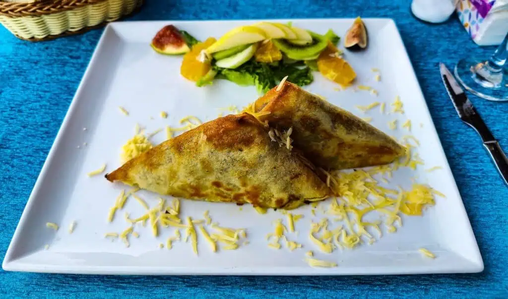 View of a vegetarian pastilla face down on a plate. It is cut into two slices and is surrounded by cheese on a white plate. There is also a salad ont he plate. You can try this during one of the best food tours in Marrakech.