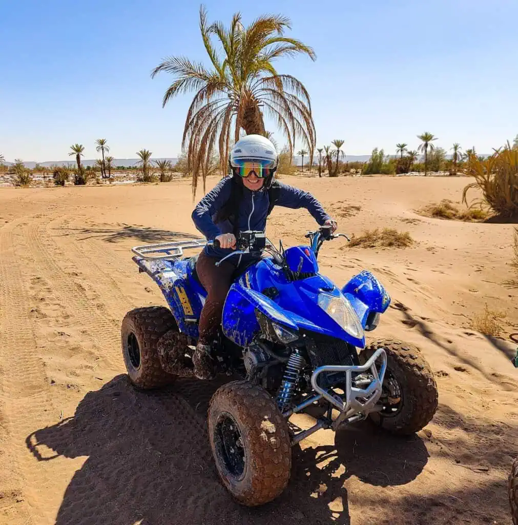 Me riding a blue quad bike through a desert with palm trees. I am waering a helmet, goggles, a long sleeve blue shirt and brown pants.