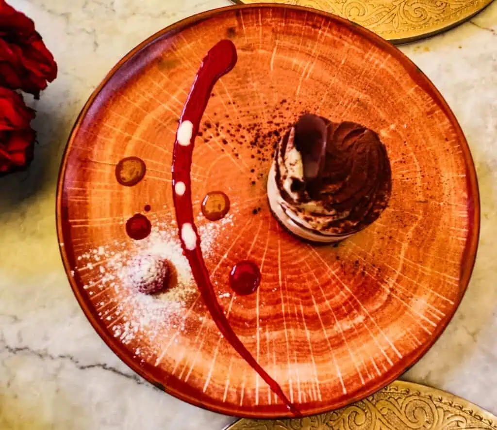 Aerial view of a tan plate with a round tiramisu from one of the most romantic restaurants in Marrakech. You can see respberry sauce beautifull presented on the side of the plate.
