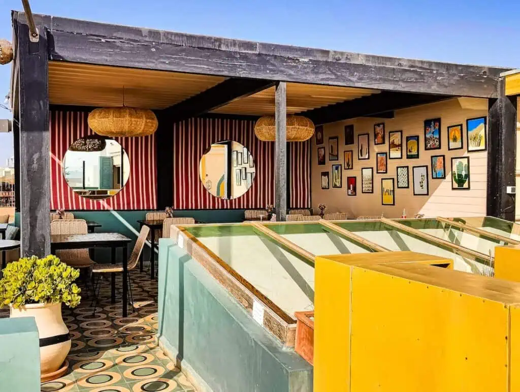 View of the sleek and stylish decor on the roof of le Palazzo in Essaouira. You can see vertical red wallpaper and round mirrors as well as hangin lamps and pictures on the wall.