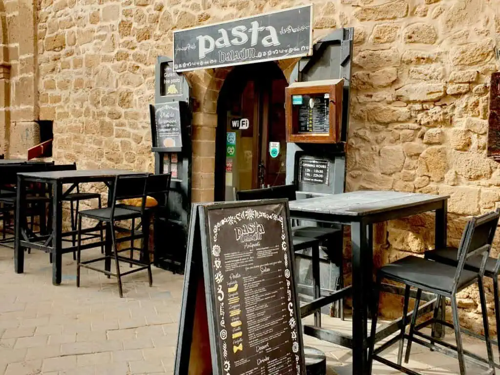 View of the black sign with white writing that says Pasta Baladin with black tables out front.