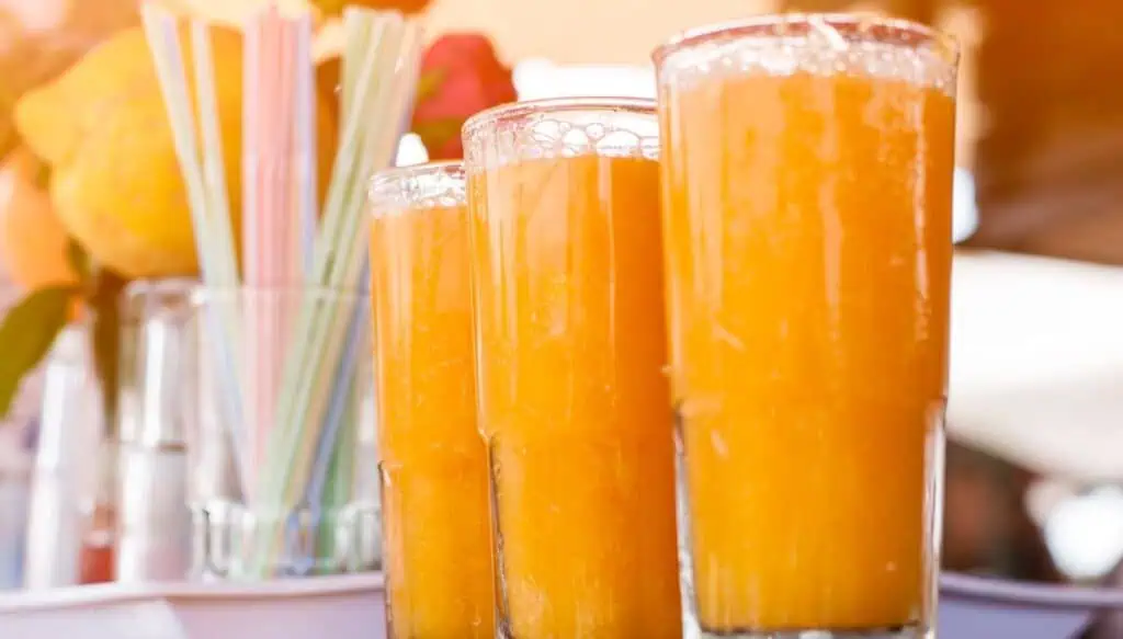 A view of three tall glasses of fresh orange juice with a glass full of straws sitting in the background.
