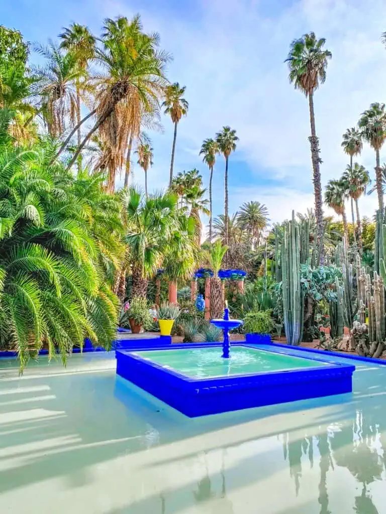 View of the incredible blue fountain inside Jardin majorelle. It is surrouned by palm trees on a sunny day. This is what you'll see when visiting Jardin Majorelle.