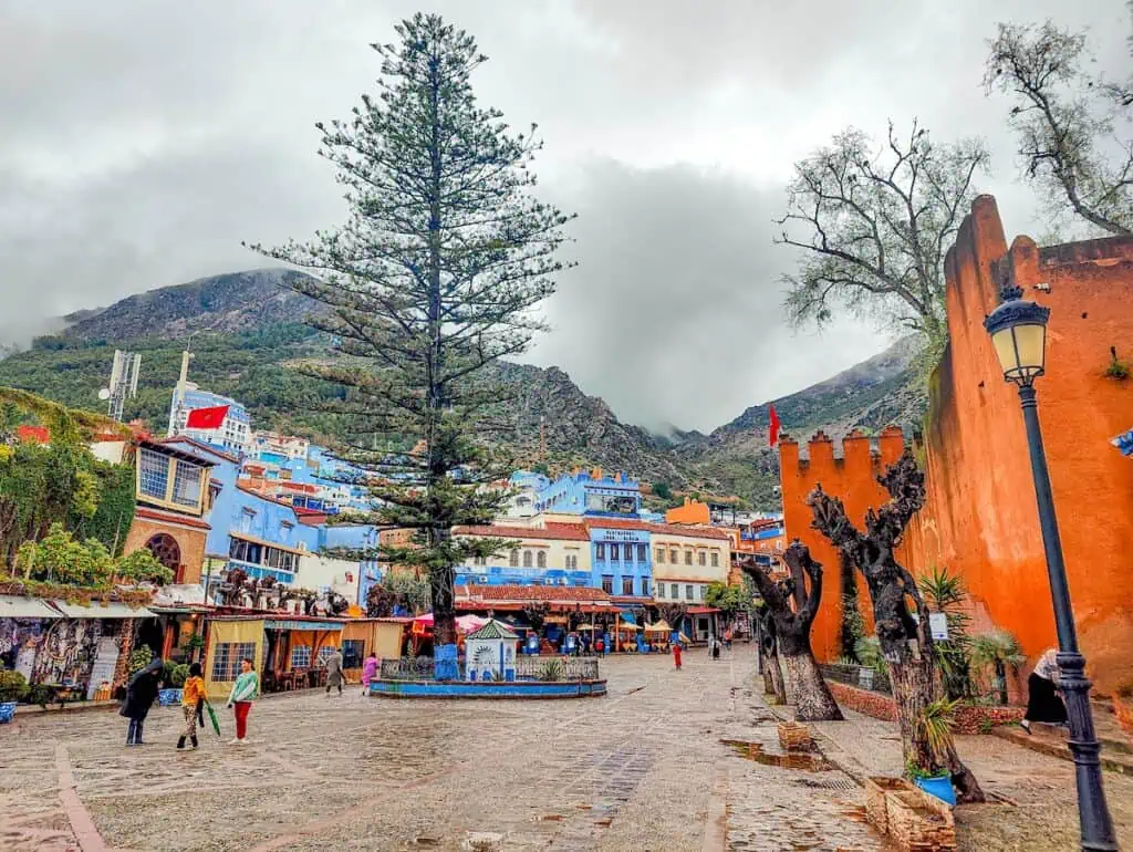 View of Plaza Uta El Hammam on a cludy day. You can see the casbah on the right and restaurants on the left with a giant pine tree in the middle.
